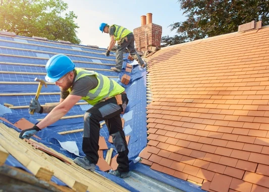 This image shows two professional roofers working on a rooftop, wearing blue hard hats, safety vests with reflective stripes, and protective gear.